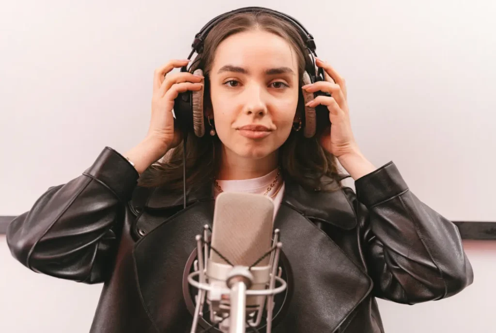 Confident young female vocalist wearing headphones and standing behind a studio microphone in a recording booth, preparing to record vocals—an example of vocal tracking that often takes place in top-tier recording spaces like Bay Eight Studio.