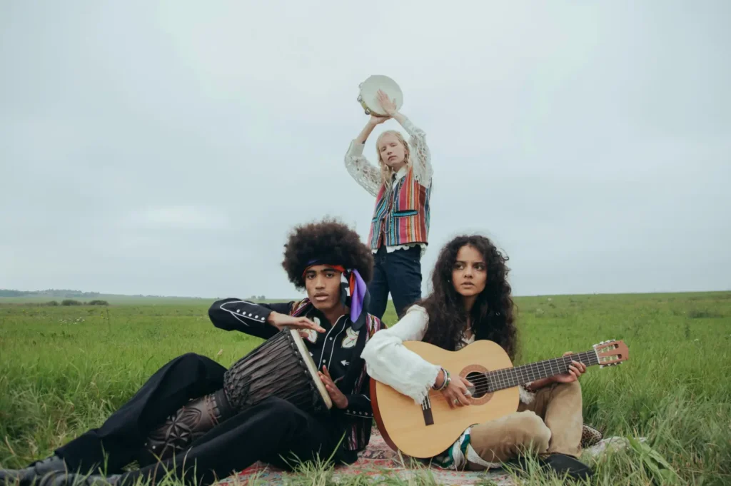 Three young musicians dressed in bohemian clothing sit in a grassy field under an overcast sky—one playing a djembe drum, another strumming an acoustic guitar, and the third holding a tambourine overhead—evoking a free-spirited vibe reminiscent of live sessions or acoustic pre-productions that might later be refined at Bay Eight Studio.