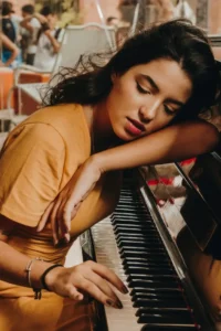 Young woman, Marcela Alessandra, in a mustard dress resting her head gently on a piano as she softly plays the keys, captured in a reflective moment of songwriting—highlighting the emotional side of music creation, which often comes to life in creative hubs like Bay Eight Studio.