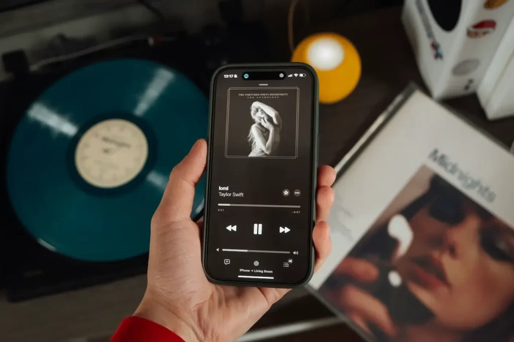 A hand holds a phone showing a playlist of Taylor Swift songs next to a teal vinyl record and an album cover on a table.