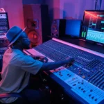 Music producer adjusting a sound mixing console under colorful studio lights, focusing on creating high-quality audio in the music production process.